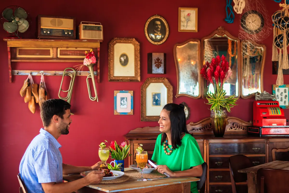 Casal sorrindo à mesa, com drinks, em restaurante colorido do Village